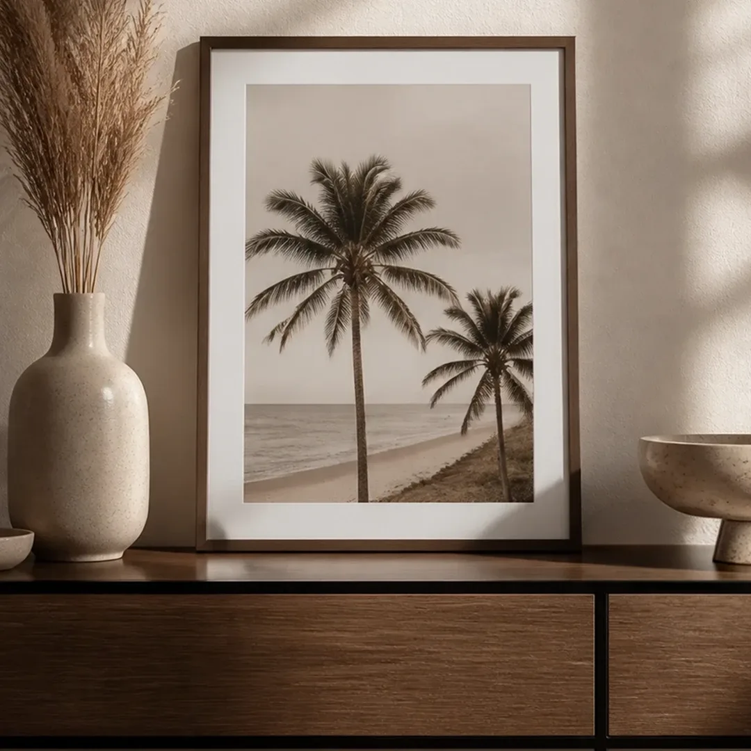 Dunkler Holzrahmen mit Palmenmotiv auf einem Holz-Sideboard, daneben Vase mit Pampasgras