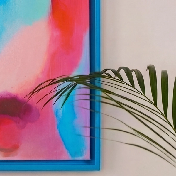 Blauer Rahmen mit buntem Bild in Pink und Türkis an einer hellbeigen Wand. Ein grünes Palmblatt ragt ins Bild hinein.