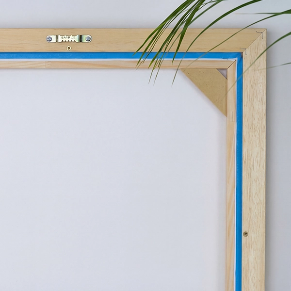 Rückansicht eines Schattenfugenrahmens mit Zackenaufhänger und Leinwand vor hellbeiger Wand. Ein Palmblatt ragt ins Bild.